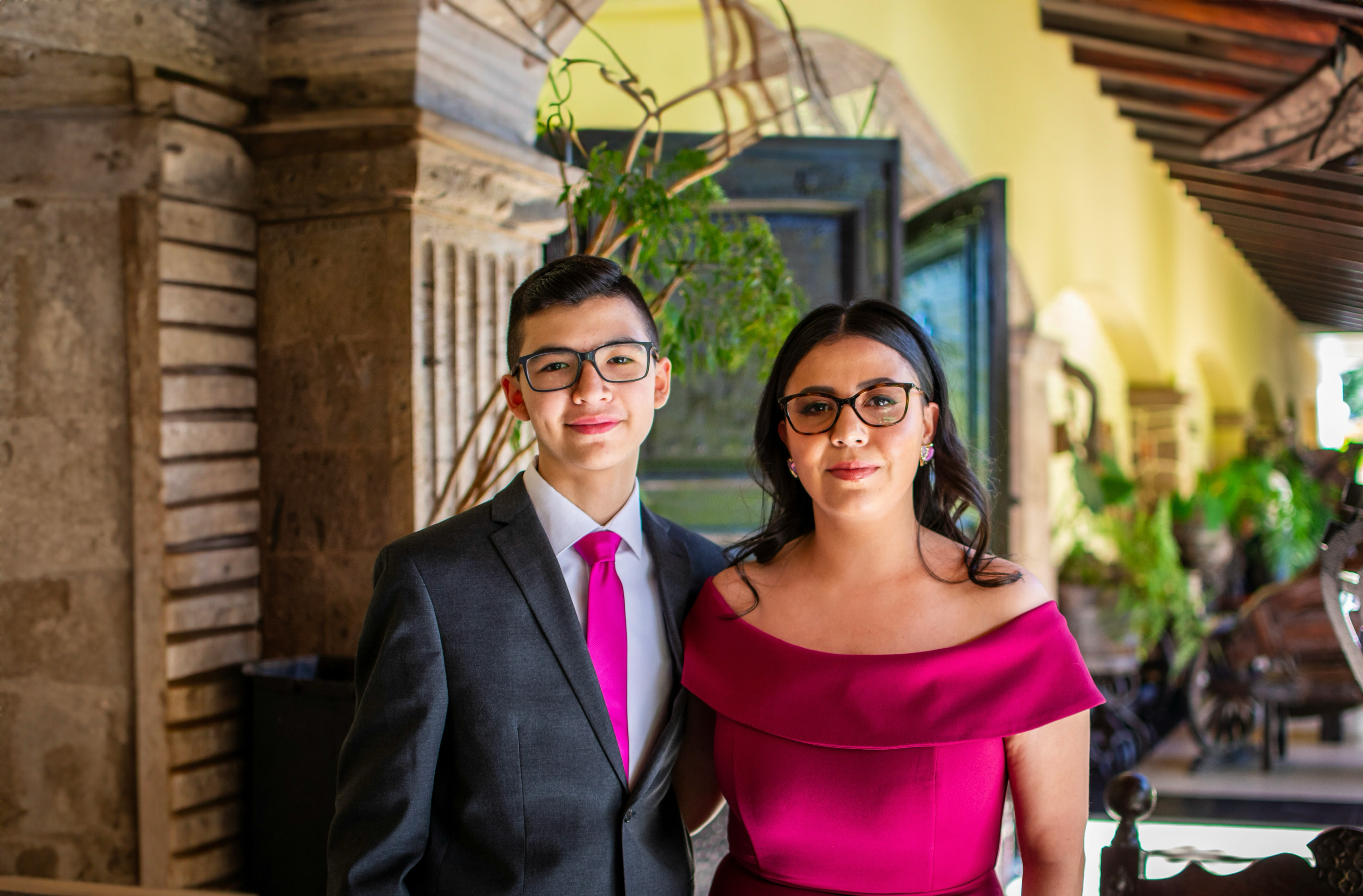 A mother and her son at a formal event in formal attire. The mother is wearing a fuschia dress while the son is in a dark-colored suit with a matching tie.