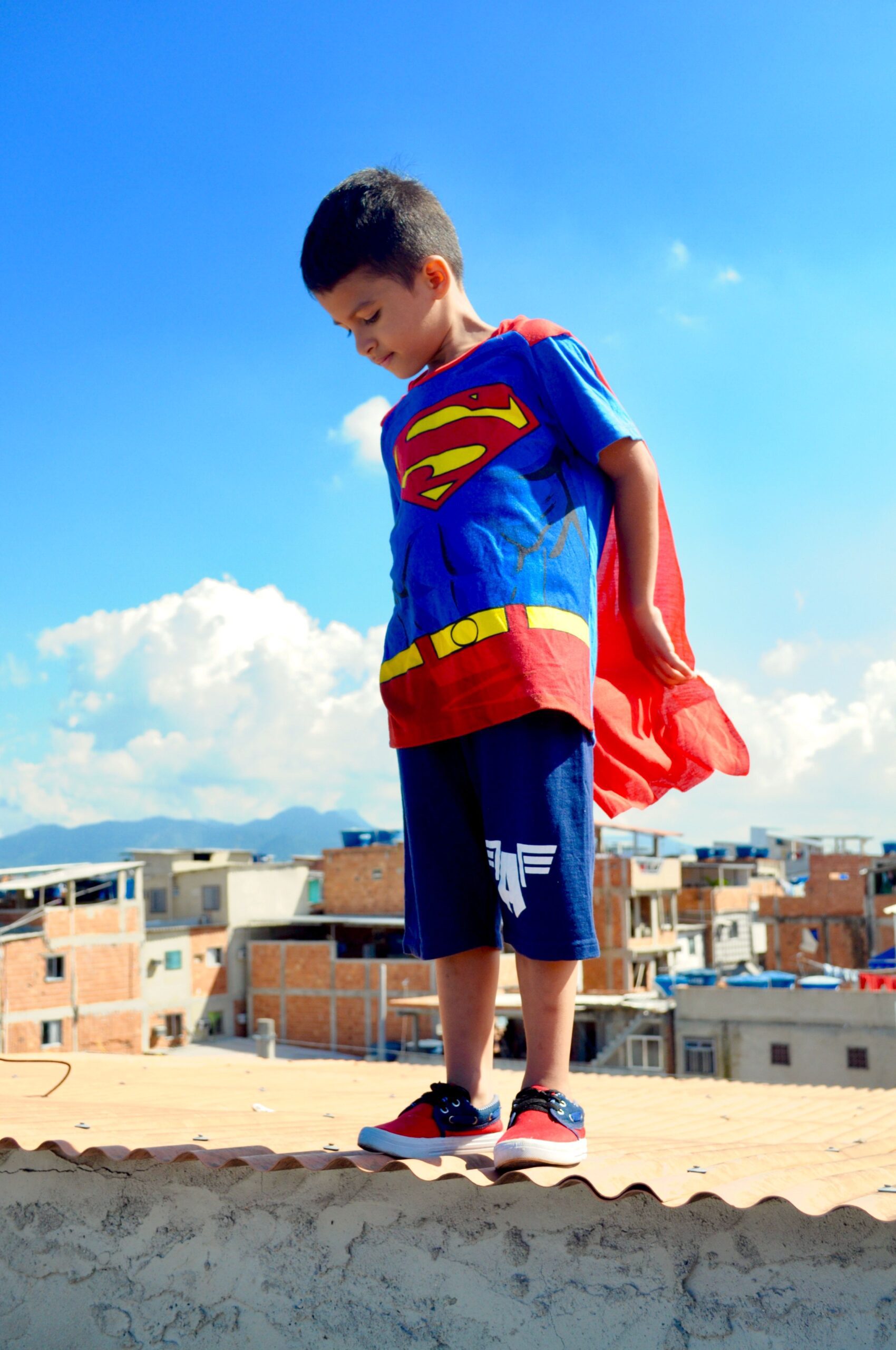 A child dressed as Superman stands on a rooftop, with buildings and blue sky in the background.